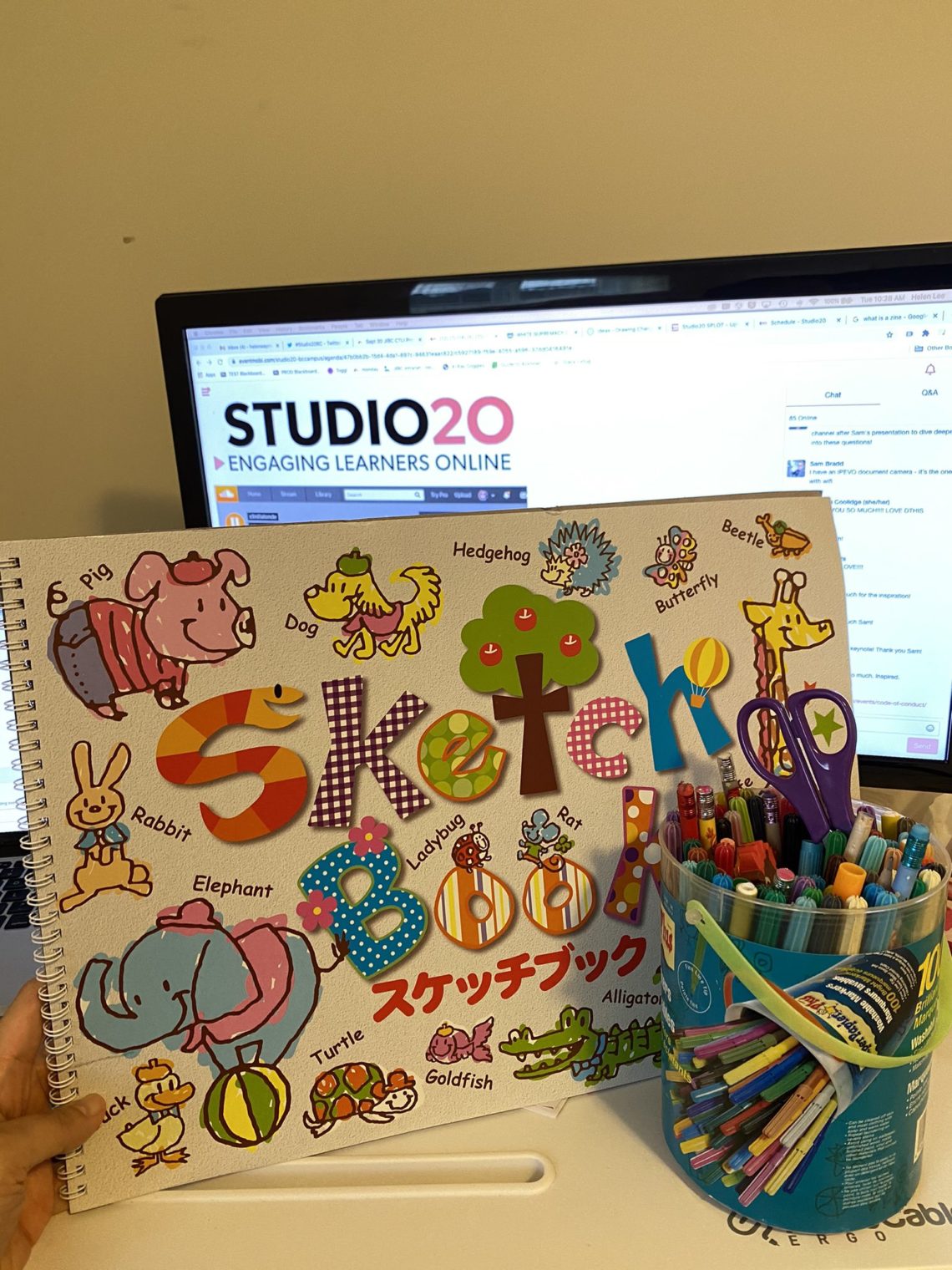 computer screen displays Studio20 conference logo, a large sketchbook pad and a pile of kids markers and a pair of kids scissors
